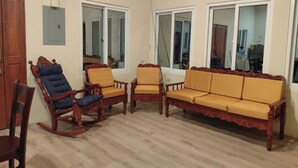 A room with a wooden floor, a rocking chair, a sofa, and two armchairs. There are windows with white frames and a door.