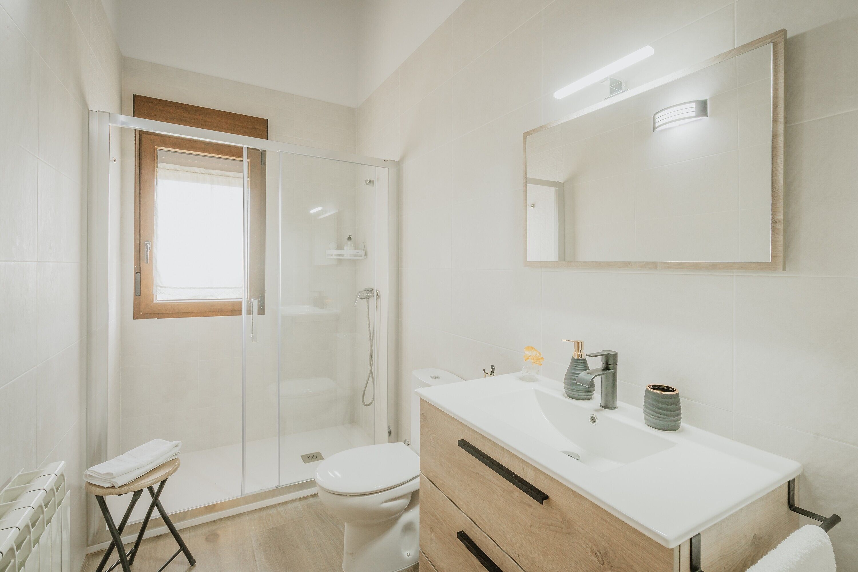 A modern bathroom with a glass shower enclosure, a toilet, a sink with a mirror, and a small stool with a towel.