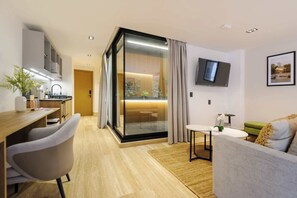 Ein modernes Wohnzimmer mit einer Couch, einem Essbereich mit Tisch und Stühlen, einer Küchenzeile und einer Glastür zum Balkon mit Pflanzen.