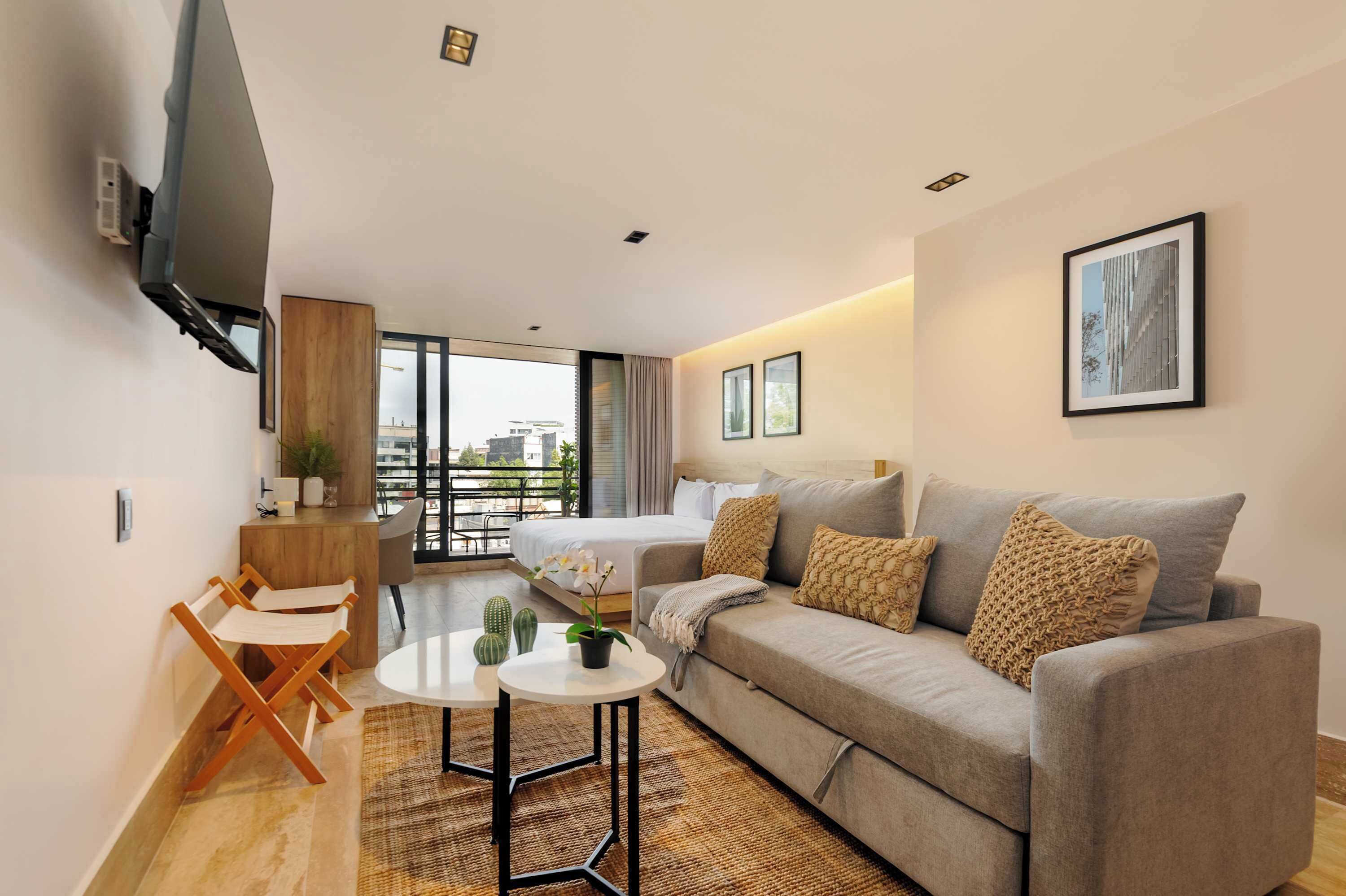 Ein modernes Hotelzimmer mit einer Couch, einem kleinen Tisch, einem an der Wand befestigten Fernseher, einem Fenster mit Blick auf Gebäude und einer Tür, die auf einen Balkon führt.
