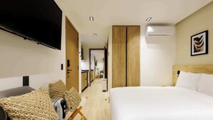 Ein Hotelzimmer mit Bett, Fernseher, Schreibtisch mit Stuhl und einer kleinen Küchenzeile im Hintergrund.