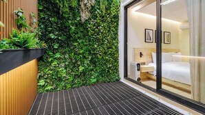 Ein modernes Hotelzimmer mit einem großen Bett, einem Nachttisch, einem Heizkörper und einer Glastür, die auf einen Balkon mit einer grünen Wand und Pflanzen führt.