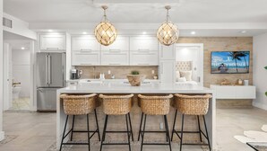 Een moderne keuken met witte kastjes, een groot eiland met barstoelen, een koelkast en een televisie waarop 'Luxury Rentals Miami Beach' staat.
