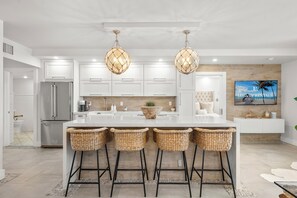 A modern kitchen with white cabinets, a large island with bar stools, a refrigerator, and a TV displaying 'Luxury Rentals Miami Beach'. - 1BR+Den Private Residence at Roney Palace (Miami Beach)