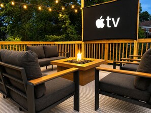 An outdoor seating area with a fire pit, a TV screen displaying the Apple TV logo, and a wooden deck. - NEW! The Dwelling Place w/ Rooftop Theater, Fire Pit, Game Room near World Cup (Atlanta)
