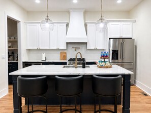 A modern kitchen with white cabinets, a central island with bar stools, stainless steel appliances, and pendant lighting. - NEW! The Dwelling Place w/ Rooftop Theater, Fire Pit, Game Room near World Cup (Atlanta)