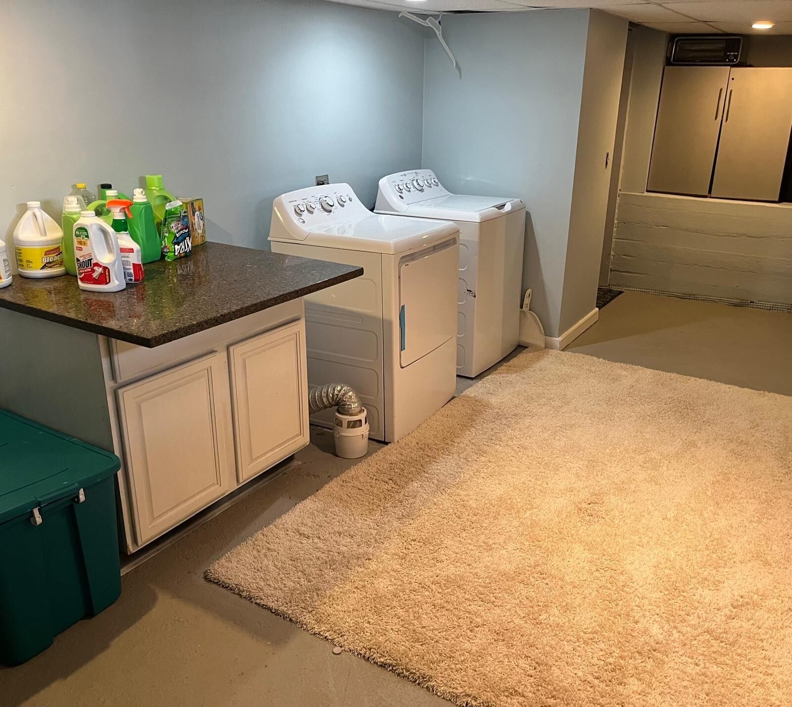 Un cuarto de lavandería con dos lavadoras, una encimera con productos de limpieza y un cesto de almacenamiento verde.