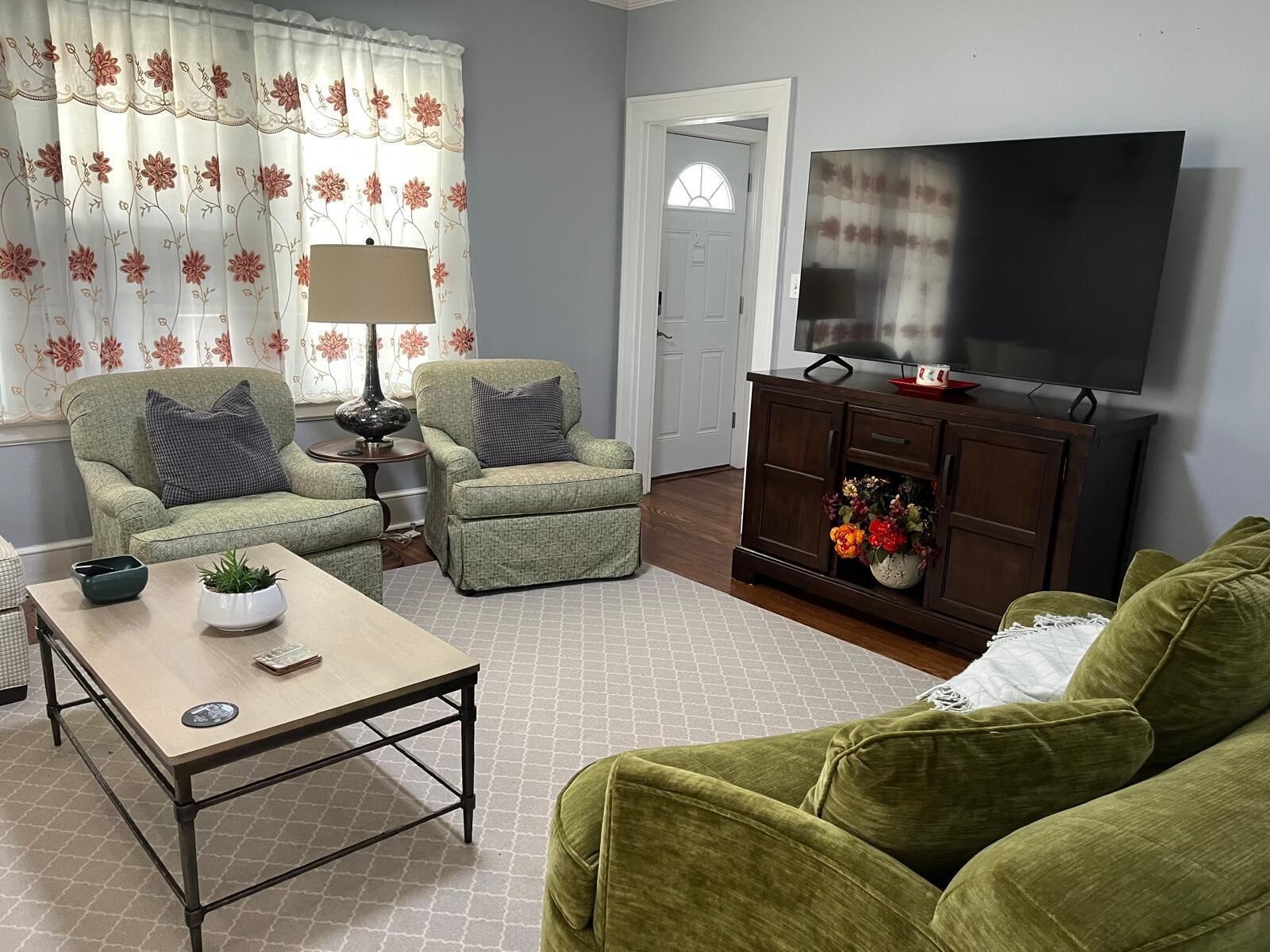 Un salon comprenant un canapé, un fauteuil, une table basse, un téléviseur et un meuble orné de fleurs.