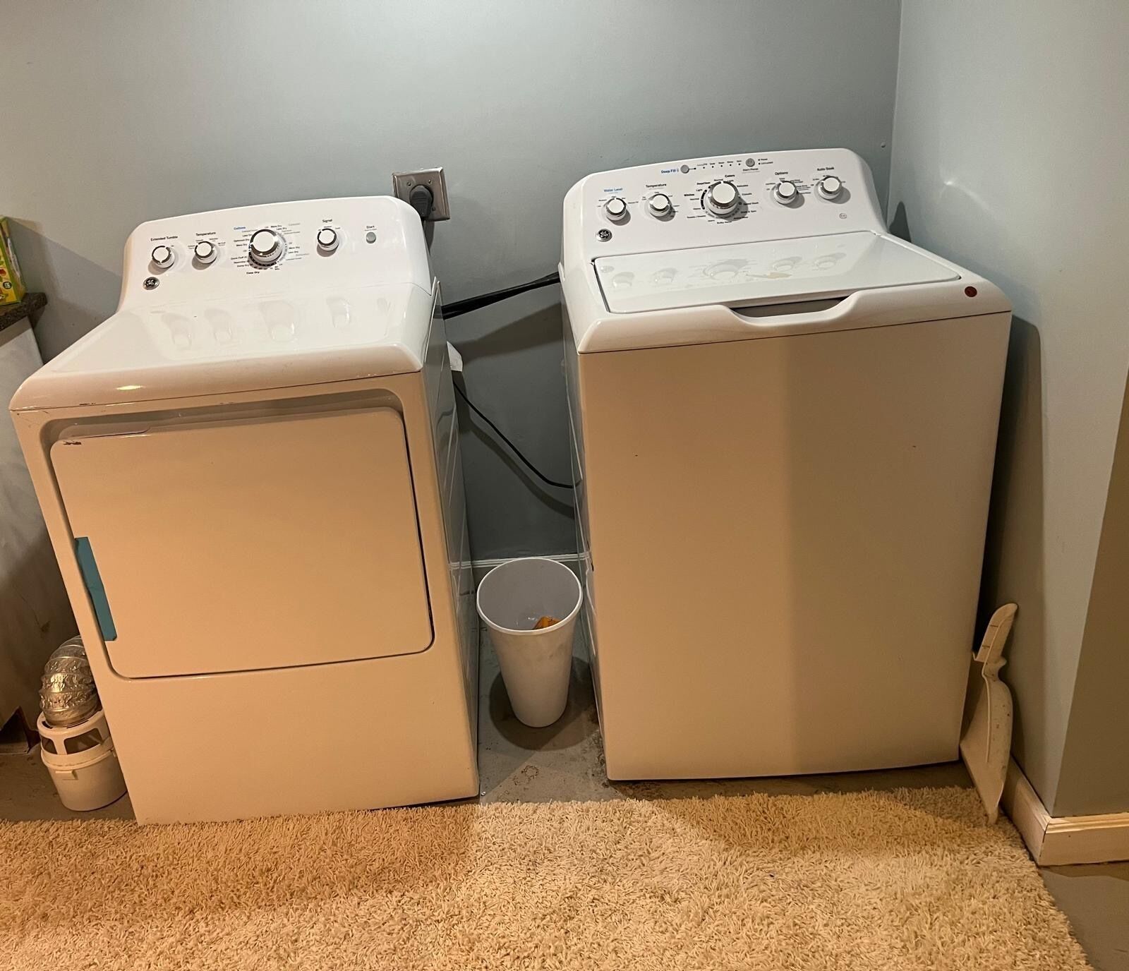 Deux machines à laver à chargement frontal, de couleur blanche, sont placées dans un coin, à côté d’une petite poubelle et d’un seau blanc.