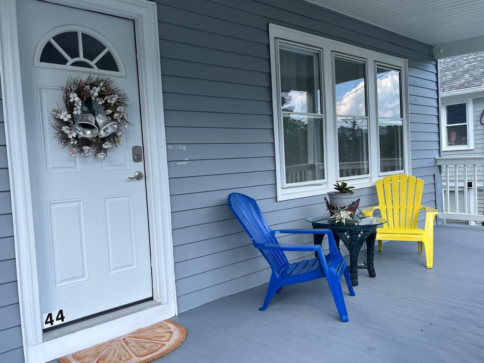 A porch with a white door, a blue and a yellow Adirondack chair, and a small table with a potted plant.