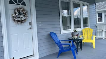 A porch with a white door, a blue and a yellow Adirondack chair, and a small table with a potted plant.