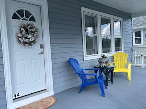Un porche con una puerta blanca, dos sillones Adirondack, uno azul y otro amarillo, y una mesita con una planta en maceta.