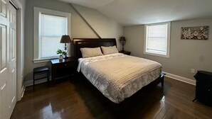 Un dormitorio con una cama, dos mesitas de noche, una lámpara de techo y una ventana con persianas.