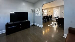 Una sala de estar moderna con una televisión de pantalla plana, piso de madera oscura, un comedor con mesa y sillas, y un pasillo que conduce a una cocina.