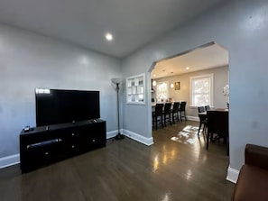 A modern living room with a flat-screen TV, dark wood flooring, a dining area with a table and chairs, and a hallway leading to a kitchen.
