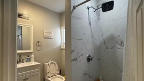 Un baño moderno con ducha, inodoro y lavamanos, paredes de mármol y una alfombrilla de ducha negra.