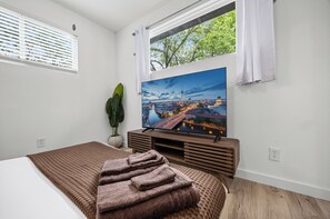 Une chambre avec une grande télévision, un lit recouvert d’une couverture marron, une plante et une fenêtre donnant sur un paysage urbain.
