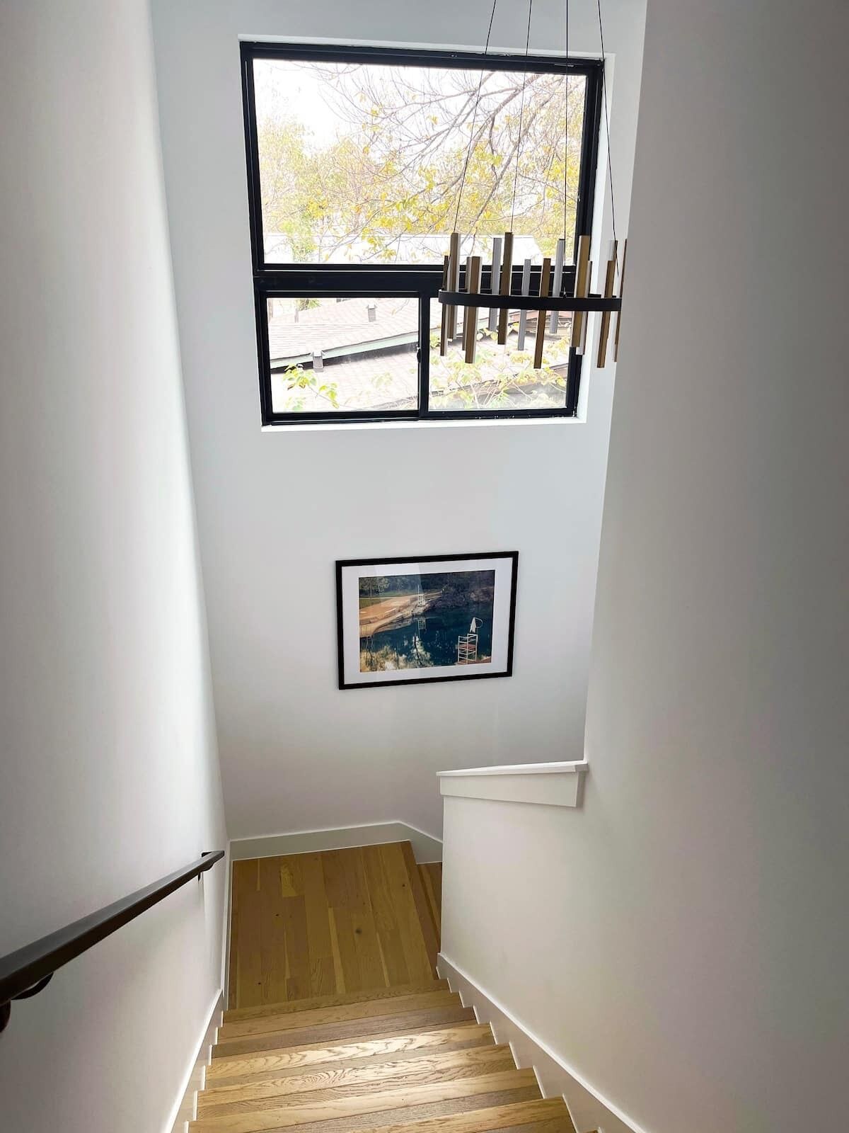 Eine Treppe mit Holzbodenstufen führt zu einer Zwischenebene, an der Wand hängt ein gerahmtes Bild und ein Fenster bietet Blick auf Bäume.