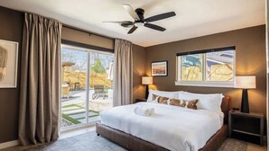 Habitación de hotel con una cama grande, ventilador de techo, dos lámparas de noche, ventana con vistas y puerta corrediza de vidrio que da a un área exterior.
