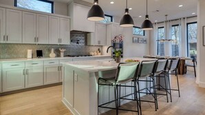 A modern kitchen with white cabinets, a central island with bar stools, pendant lighting, and a large window. - Amenities! Pool, Hot Tub, Theater, Outdoor kitchen (Austin)