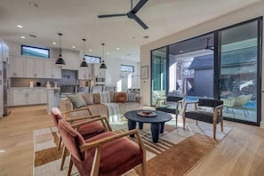 A spacious living area with a sofa, chairs, a coffee table, and a dining table. There is a kitchen in the background, a sliding glass door leading to an outdoor area, and a ceiling fan.
