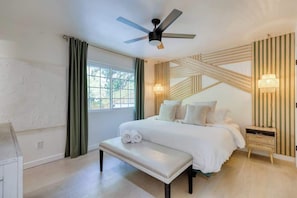 Ein Schlafzimmer mit Bett, einer Bank, einem Nachttisch, einem Deckenventilator und einem Fenster mit Vorhängen.