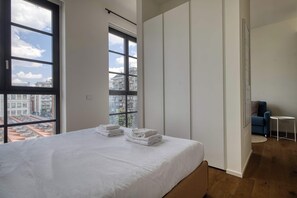 A bedroom with a large window, a bed with neatly folded white towels, a blue chair, and a small white table. - w* | Bright 1BR near to the center of Milan (Milano)