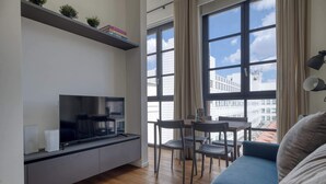 Ein modernes Wohnzimmer mit Flachbildfernseher, einer Couch, einem Esstisch mit Stühlen und großen Fenstern mit Vorhängen.