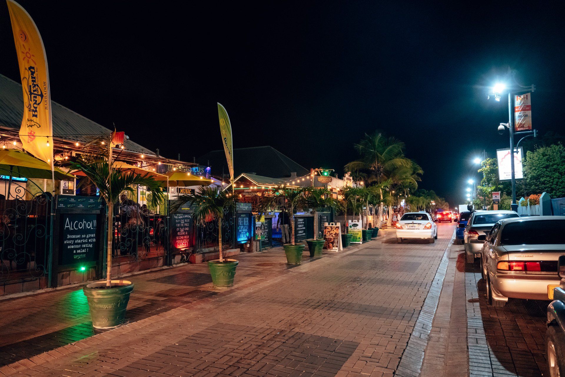 Escena nocturna de una calle con mesas al aire libre, palmeras y coches estacionados a lo largo de la vía.