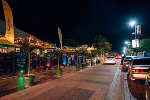 Escena nocturna de una calle con mesas al aire libre, palmeras y coches estacionados a lo largo de la vía.