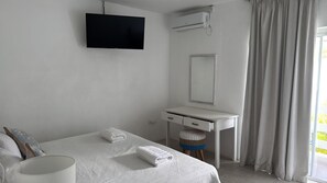 Una habitación de hotel con cama, un escritorio blanco, un televisor de pantalla plana, un espejo y una ventana con cortinas.