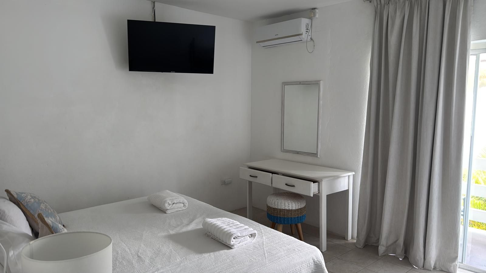 A hotel room with a bed, a white desk, a flat-screen TV, a mirror, and a window with curtains.