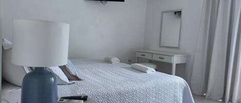 A hotel room with a bed, a desk with a lamp, a television, a mirror, and an air conditioning unit.