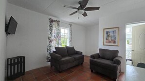Una habitación con dos sofás, un ventilador de techo, una ventana con cortinas, una puerta y un televisor montado en la pared.