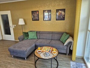 A living room with a gray sectional sofa, a coffee table with board games and snacks, a lamp, and three framed artworks on the wall.