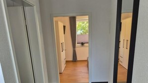 Un corridoio con un tappeto verde, pareti bianche e pavimento in legno. Si vedono porte che conducono a una stanza con finestra e tende.