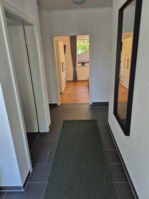 Un pasillo con un felpudo verde, paredes blancas y suelo de madera. Se observan puertas que dan a una habitación con ventana y cortinas.