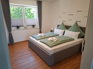 Una cama doble con ropa de cama verde y blanca, una mesita de noche con una lámpara y una ventana con cortinas.