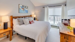 A bedroom with a bed, two bedside tables, a window with curtains, a wall-mounted sign, and a view of the outside. - Stylish and Modern Condo at Loon! Pool & Hot Tub (Lincoln)