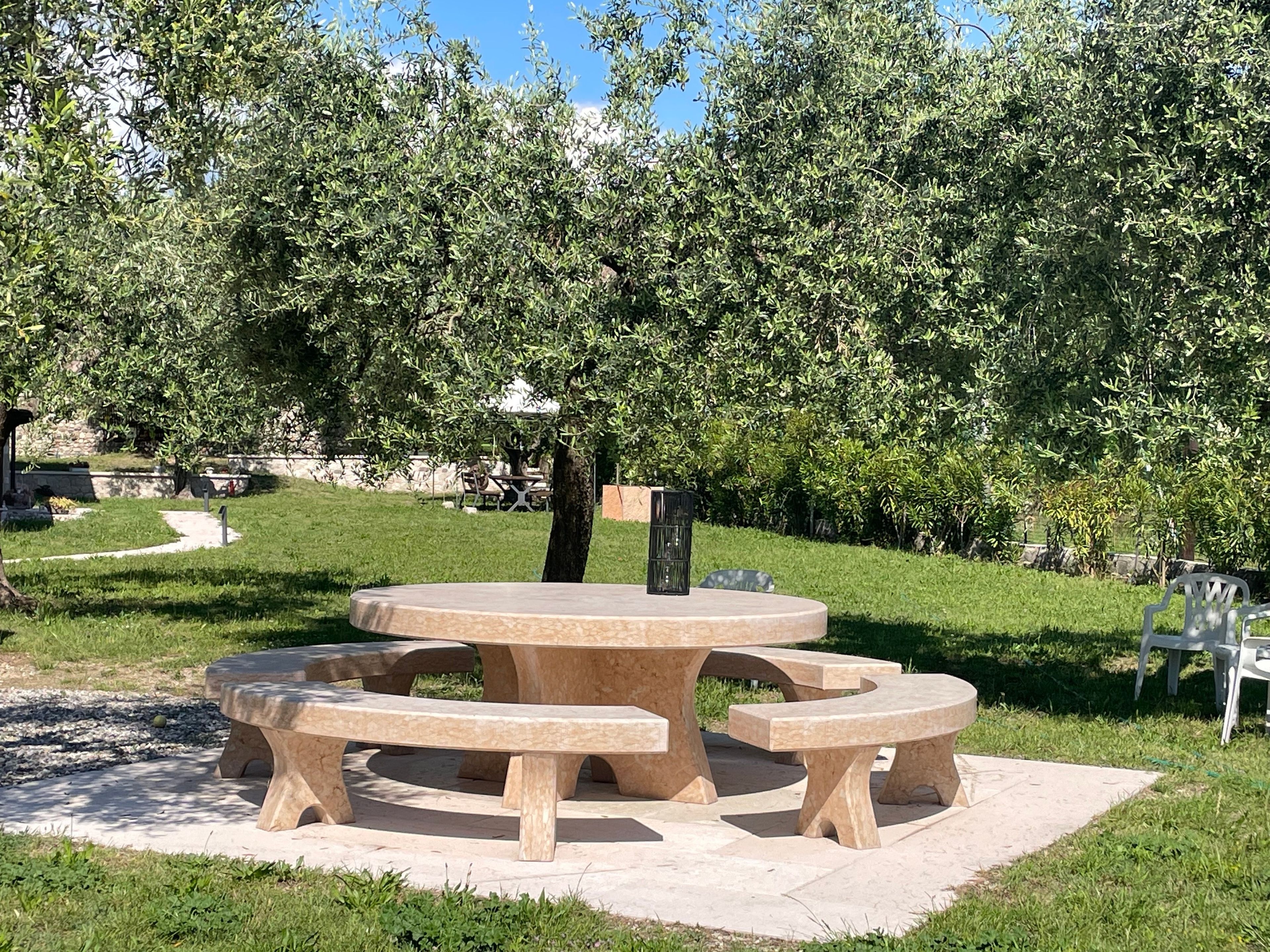 A circular stone picnic table with benches in a park with trees and a stone wall in the background.