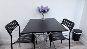 Une pièce avec une table noire, deux chaises noires, un vase avec des fleurs violettes et une poubelle noire.