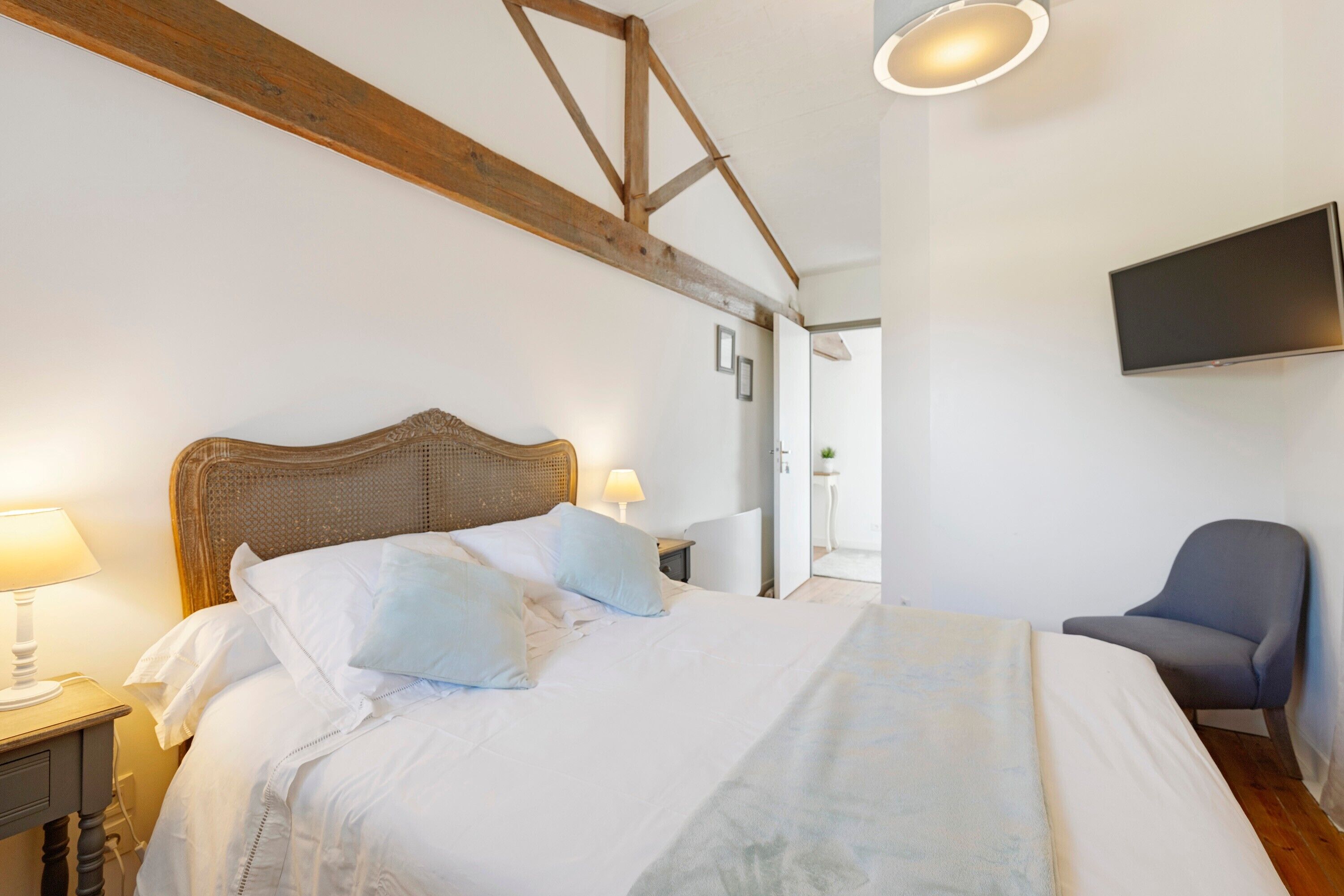 A bedroom with a wooden headboard, a bed with white bedding, a bedside table with a lamp, a chair, and a TV mounted on the wall.