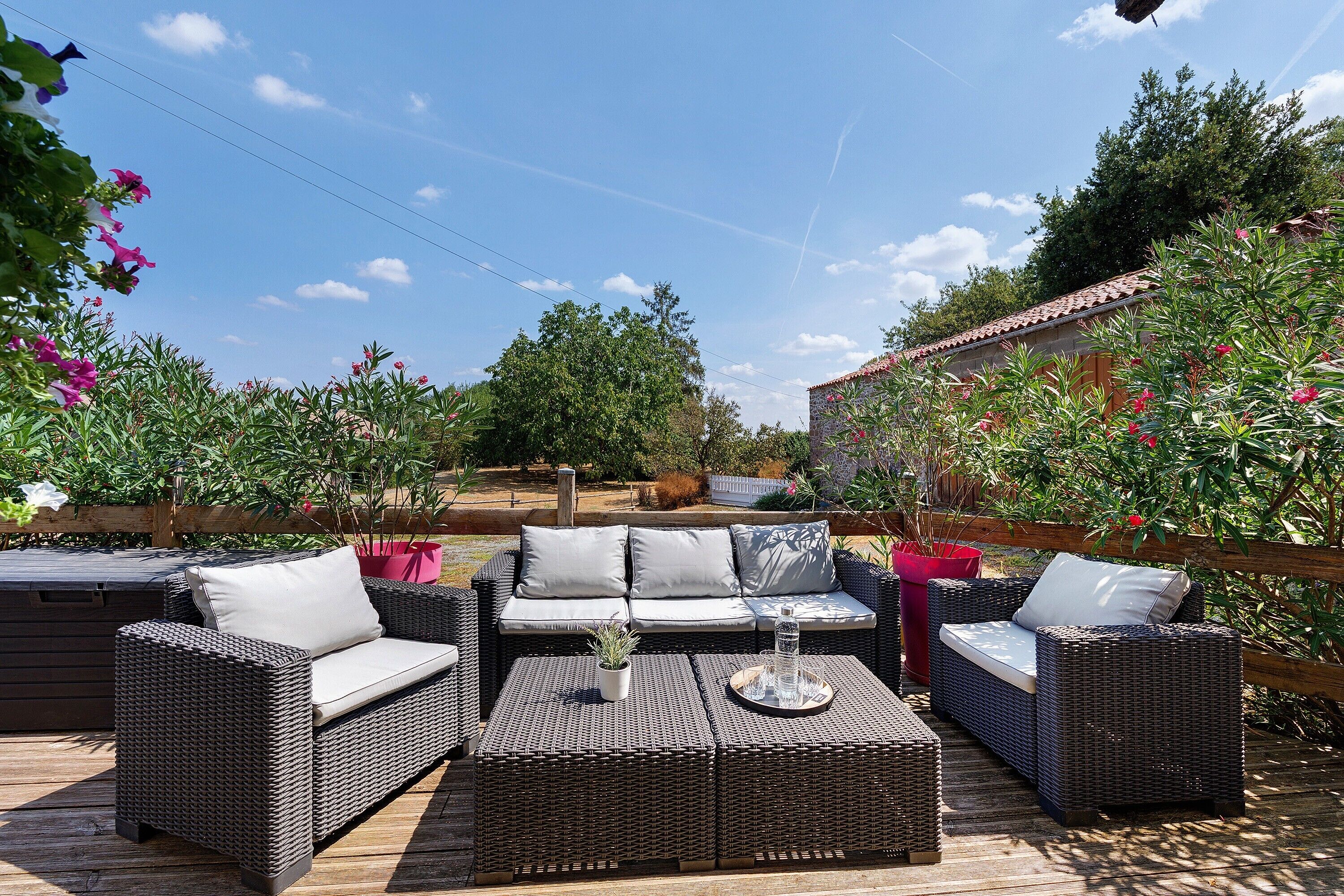 An outdoor seating area with wicker furniture and cushions, surrounded by lush greenery and flowering plants.