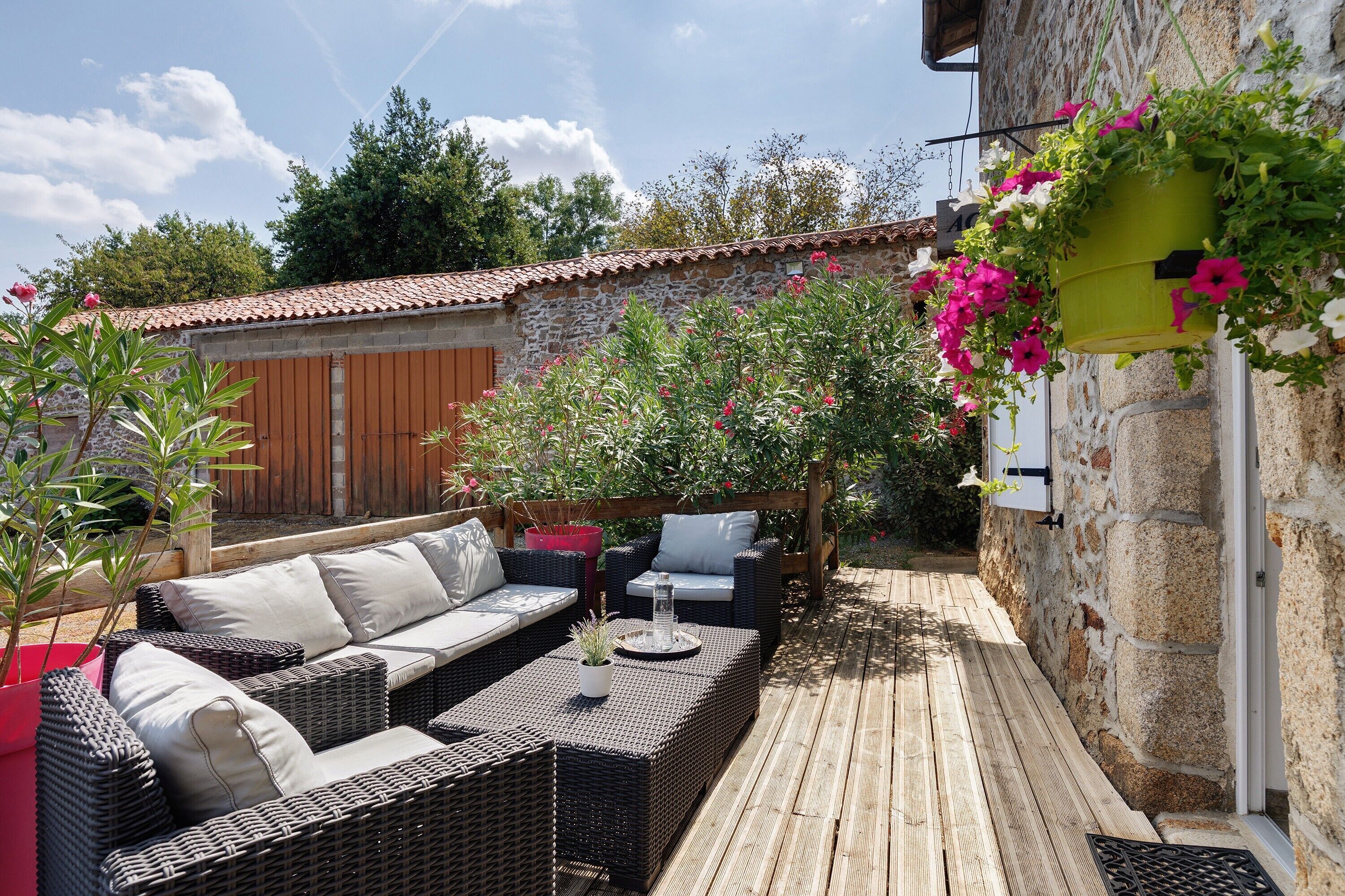 Une terrasse dotée de meubles en osier, d’un mur en pierre et de plantes en fleurs.