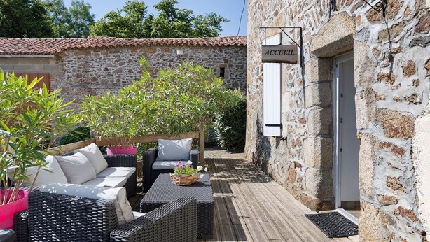 Un edificio de piedra con un letrero que dice 'ACCUEIL' y una terraza de madera con muebles de mimbre y plantas en macetas.