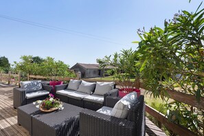 An outdoor patio with wicker furniture, a wooden deck, and a house in the background. - Eugénie - Aux Charmes d’Antan Guest Room with Shared Pool, Terrace & Wi-Fi (Sèvremont)
