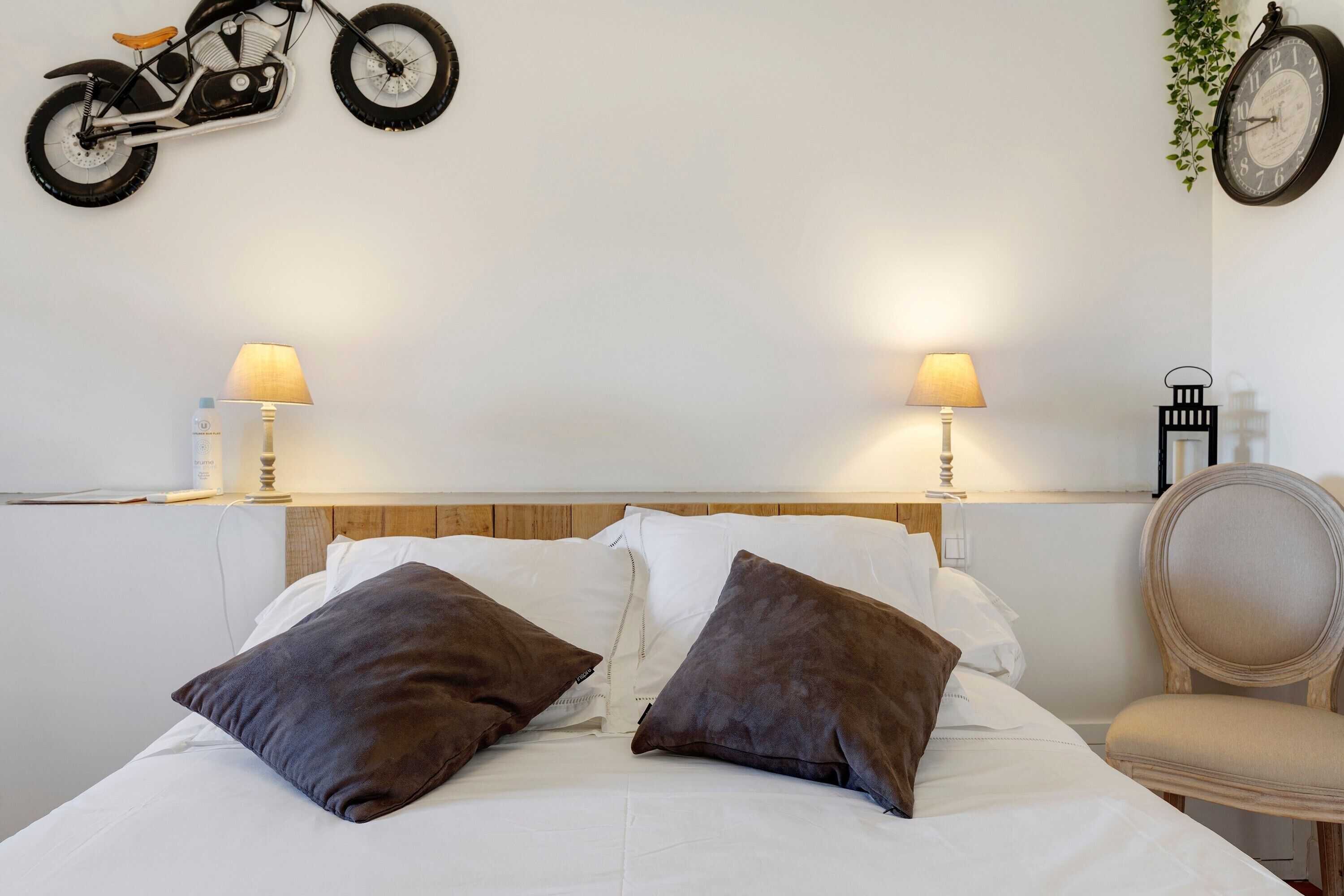 A bed with white linens and two brown pillows, a wooden headboard, bedside lamps, a wall-mounted clock, and a motorcycle decoration.