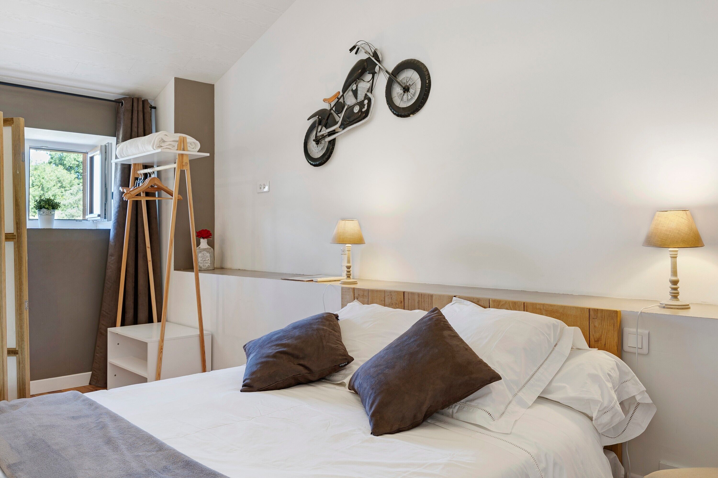 A bedroom with a bed, pillows, a wall-mounted motorcycle, bedside lamps, and a window with curtains.