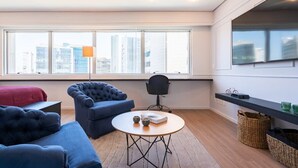 A modern living room with a large window, a round table, two blue sofas, a black chair, and a bookshelf. - #RD1011 | Cyclinn Vila Olímpia (Vila Olímpia)