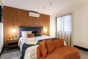 A hotel room with a wooden panel wall, a bed with orange and white bedding, a nightstand with a lamp, and a window with curtains. - #TR1109 | Cyclinn Faria Lima Itaim (Itaim Bibi)